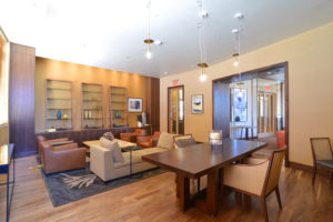 A modern lounge area in Navy Yard apartments featuring brown leather chairs, a beige sectional sofa, a wooden table, a patterned rug, bookshelves, and hanging light fixtures under a ceiling with recessed lighting.