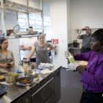 Chef in a purple jacket demonstrates a cooking technique to a group of attentive observers in a kitchen setting.