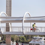 A person in a yellow vest and helmet rides a bicycle across a modern arched footbridge above the marina's intricate floor plan of boats.