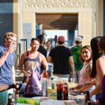 A group of people at an outdoor gathering, standing around a table with drinks and snacks. Some are talking, others are preparing food. Bright daylight.