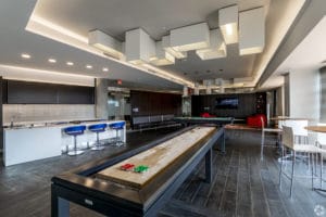 A modern recreational room with a shuffleboard table, bar seating, and contemporary lighting fixtures.