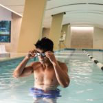 Man standing in an indoor swimming pool adjusting his goggles, near a lane divider, with white modern architecture in the background.