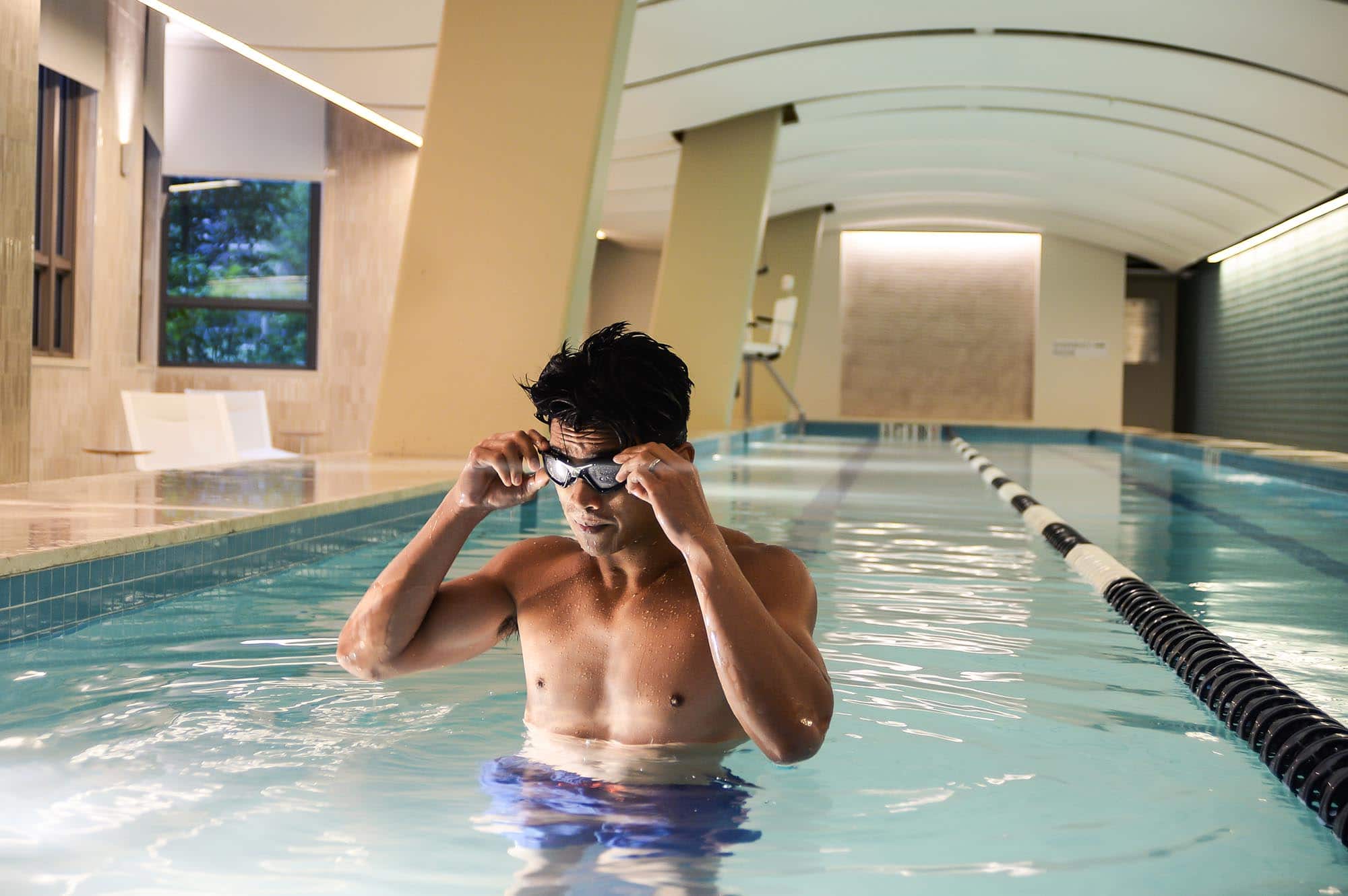 Man standing in an indoor swimming pool adjusting his goggles, near a lane divider, with white modern architecture in the background.