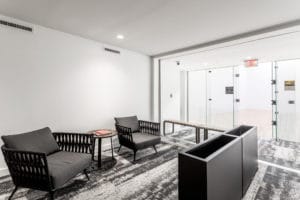 A modern waiting area with two black wicker chairs and a small table on a patterned carpet. Glass doors with exit signage are in the background.