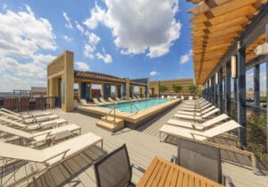 The rooftop pool, with its surrounding loungers under a partly cloudy sky, offers a serene escape. Beside the water, modern wooden structures provide ample shade for relaxation by the pool.