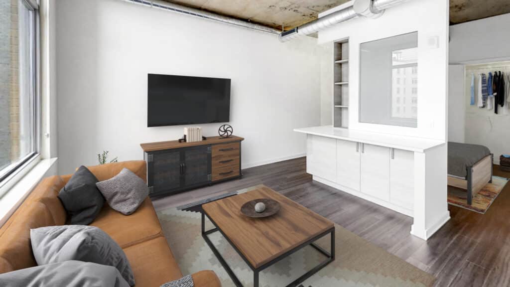 Modern living room with a large wall-mounted TV, a brown leather sofa, a wooden coffee table, and a sideboard. The adjacent bedroom is partially visible. The ceiling features exposed ductwork.