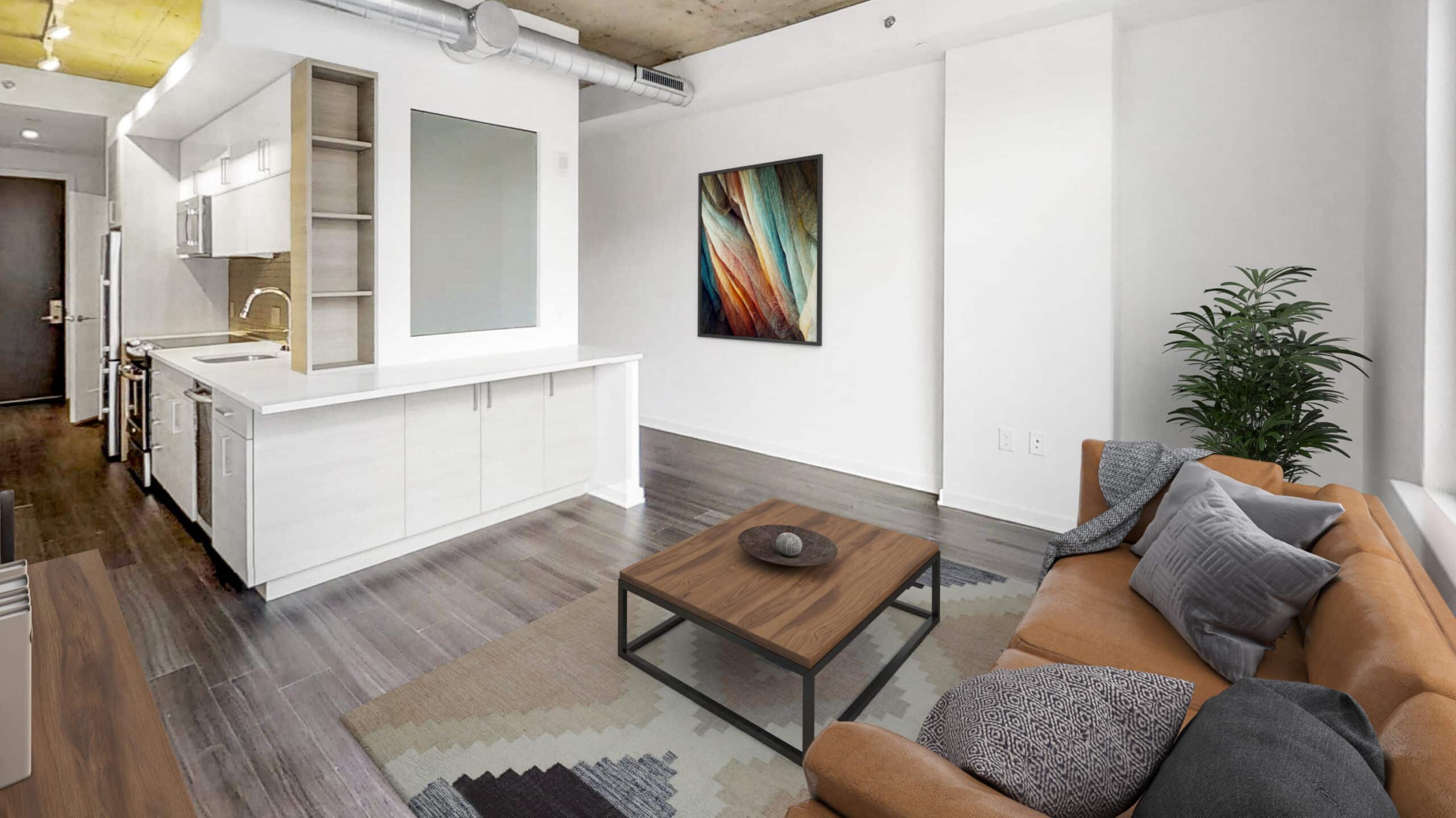 A modern, functional open-concept living area with a kitchen features wall-mounted artwork, a brown sofa with cushions, a coffee table, a rug, and a potted plant. The space has gray flooring and white walls.