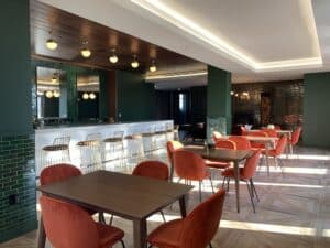 A modern lounge with a bar, green tiled walls, wooden ceiling, and tables with orange chairs. Bright lighting from ceiling fixtures.