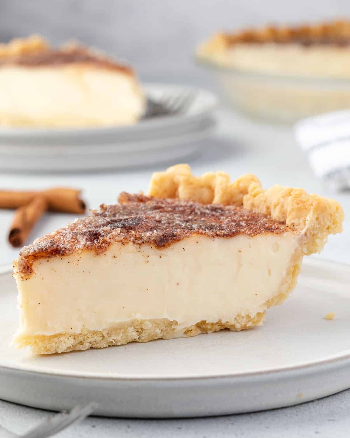 Slice of custard pie with a cinnamon-dusted top on a white plate, with a whole pie and a cinnamon stick in the background.