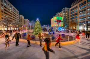 People ice skating around a decorated holiday tree in an outdoor rink surrounded by buildings with festive holiday lights at canal park in the capitol riverfront neighborhood.