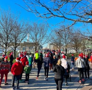 A group of people wearing Santa hats are participating in an outdoor event on a sunny day, running along a pathway lined with leafless trees.