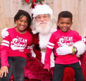 Two children in "Team Santa" shirts sit beside a man dressed as Santa Claus, all smiling.