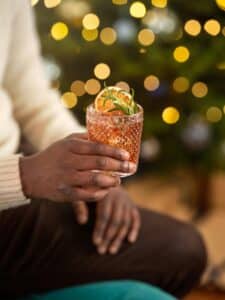 A person holding a textured glass with a festive red drink, garnished with citrus slices and herbs. Blurred holiday lights and a tree are in the background.