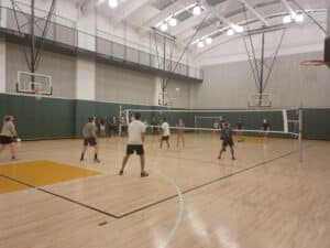 A group of people playing volleyball in a gymnasium, with two volleyball nets and basketball hoops visible in the background.