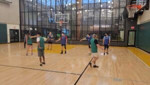 A group of people wearing green and blue jerseys play basketball on an indoor court with large windows and overhead lights.