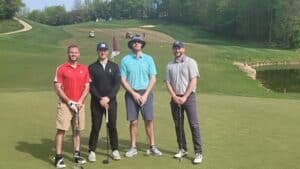 Four men stand on a golf green posing for a photo, holding golf clubs, with a golf course and golf carts visible in the background.