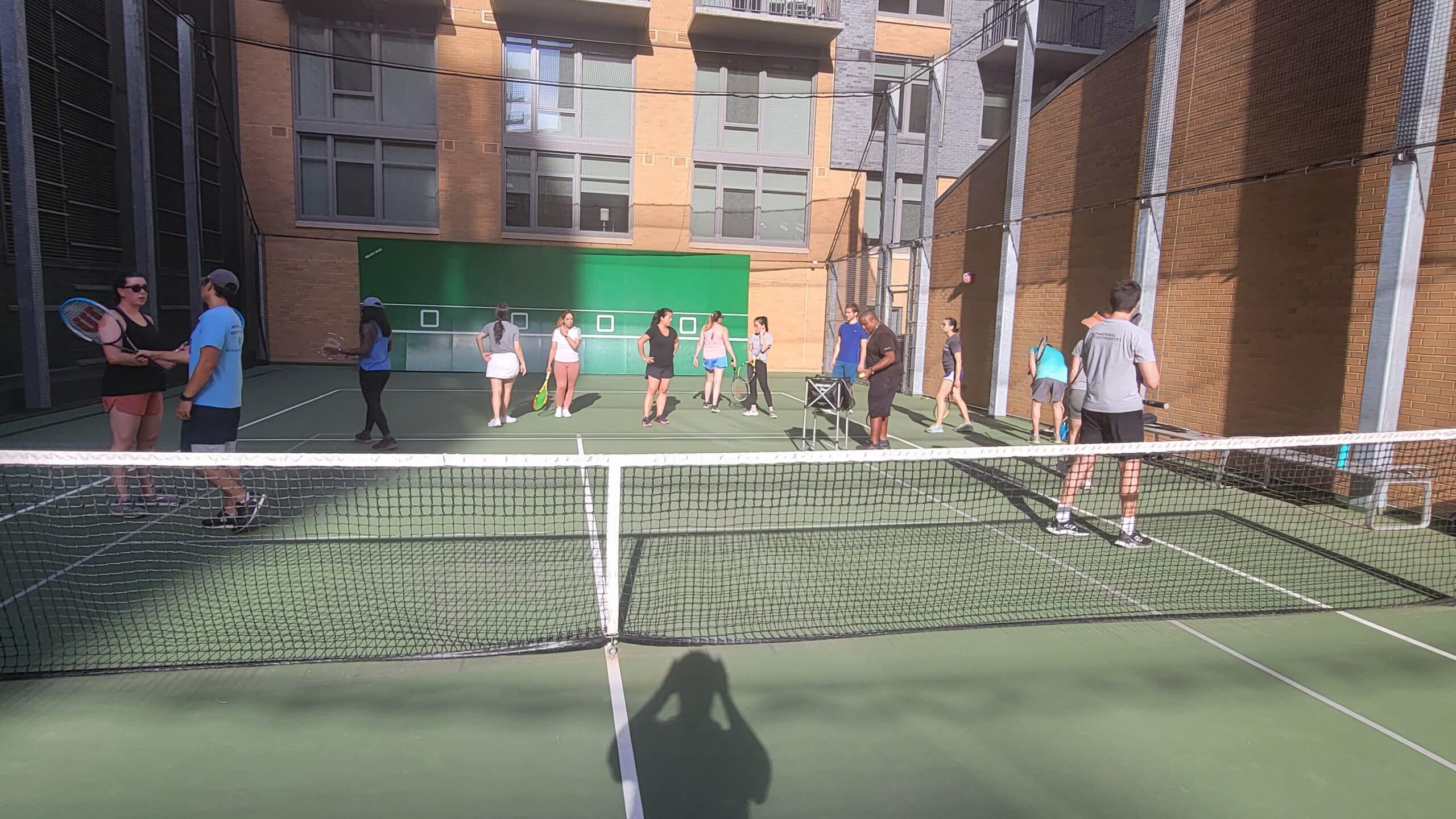 A group of people stand on an outdoor tennis court near a green wall, some holding rackets, with buildings in the background.