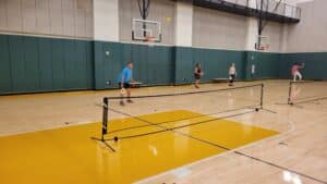 Four people play pickleball in a gymnasium with a yellow and wood floor; two on each side of a portable net, with a basketball hoop in the background.