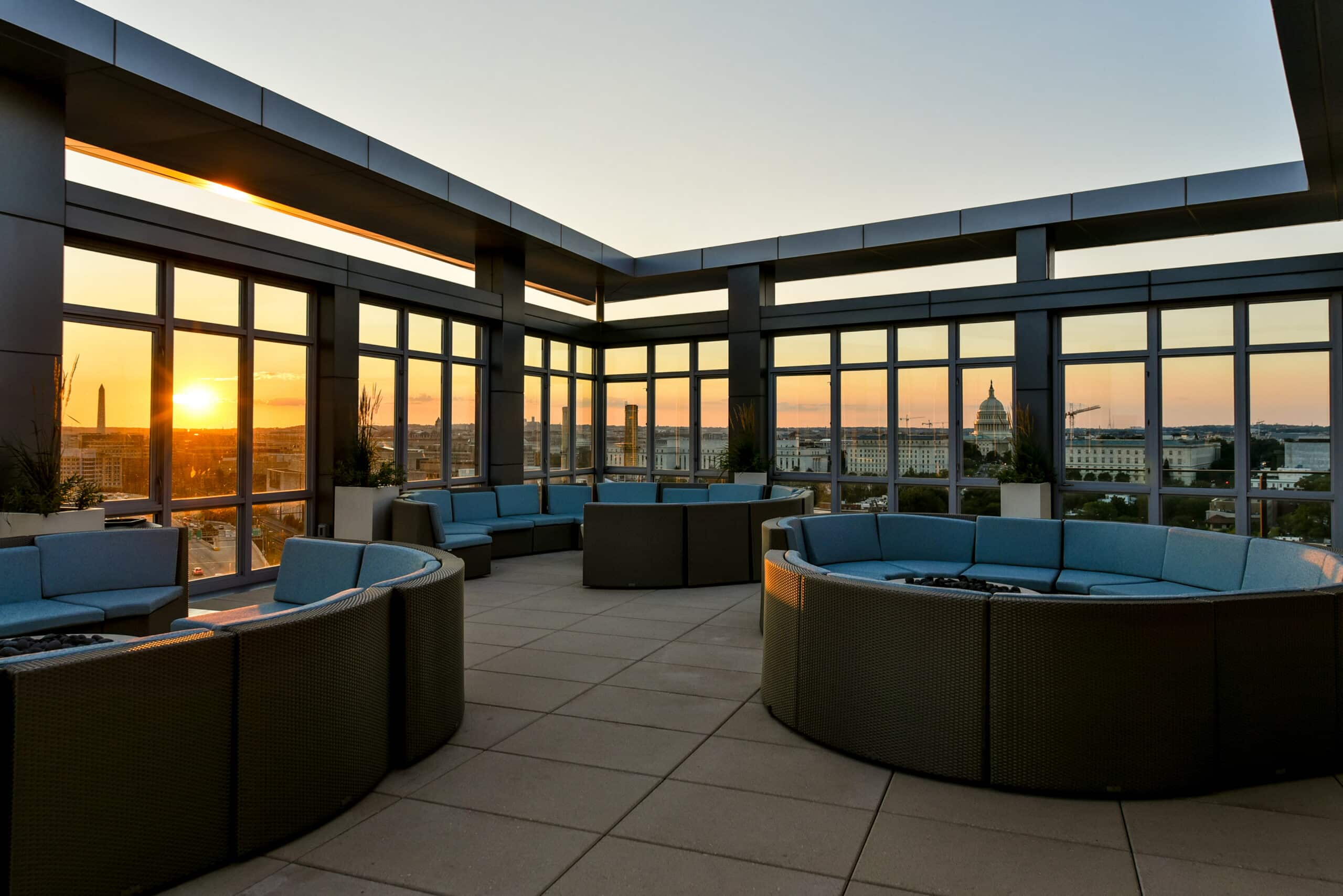 Rooftop terrace amenity with blue cushioned seating and fire pits, overlooking a city skyline at sunset, featuring large windows, modern architecture, and exceptional amenities.